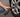 Close-up of a person kneeling on asphalt while inflating a car tire using a black portable cordless tire inflator with a digital pressure gauge display.